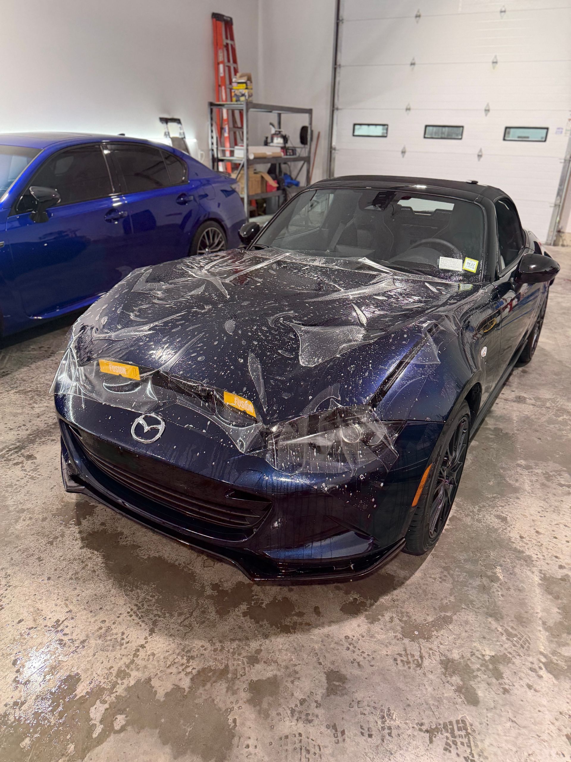 Black convertible covered in white foam in a car wash bay, with a blue car beside it.