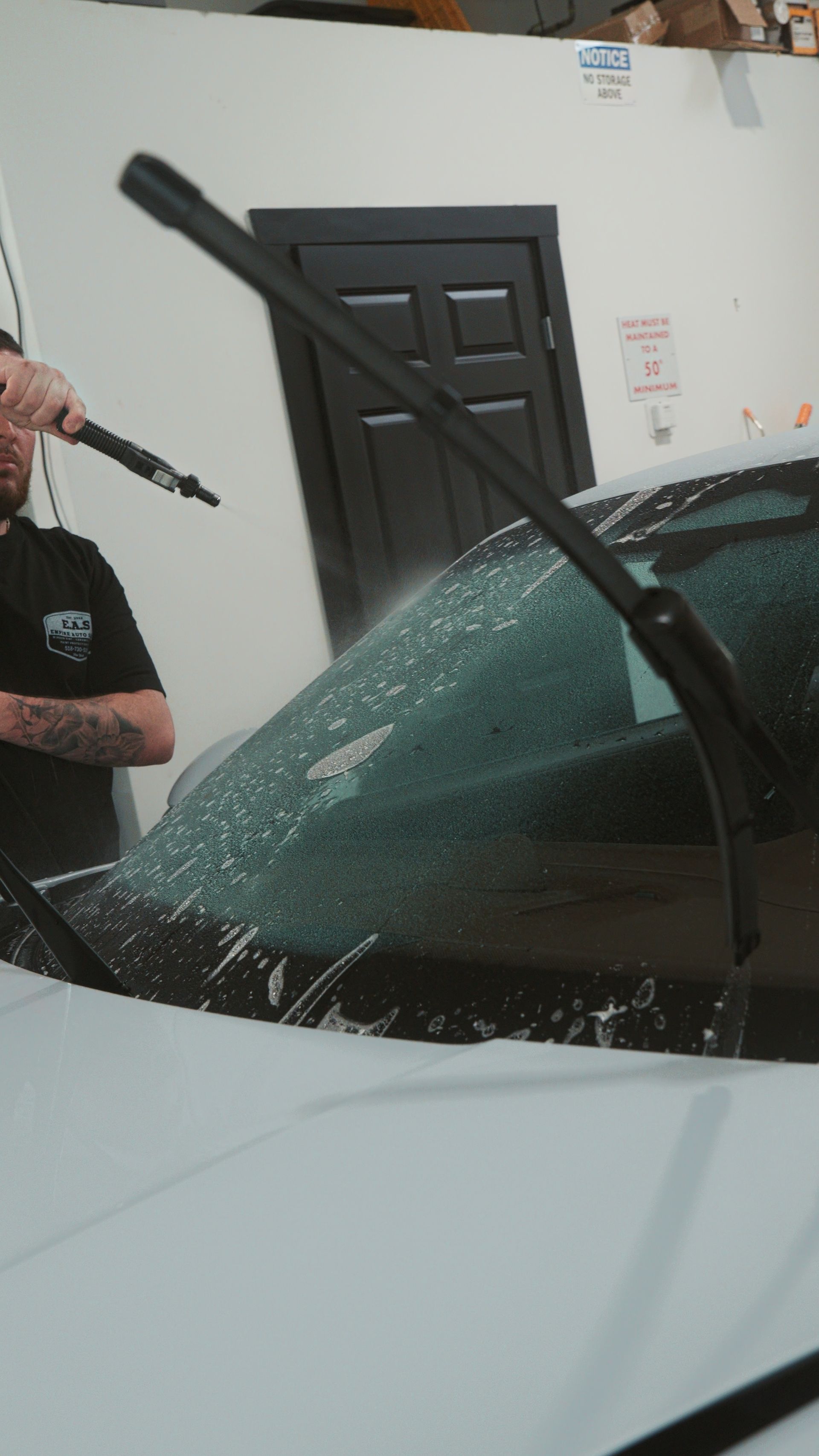 Person uses a pressure washer on a car windshield, spraying water in front of a garage door.