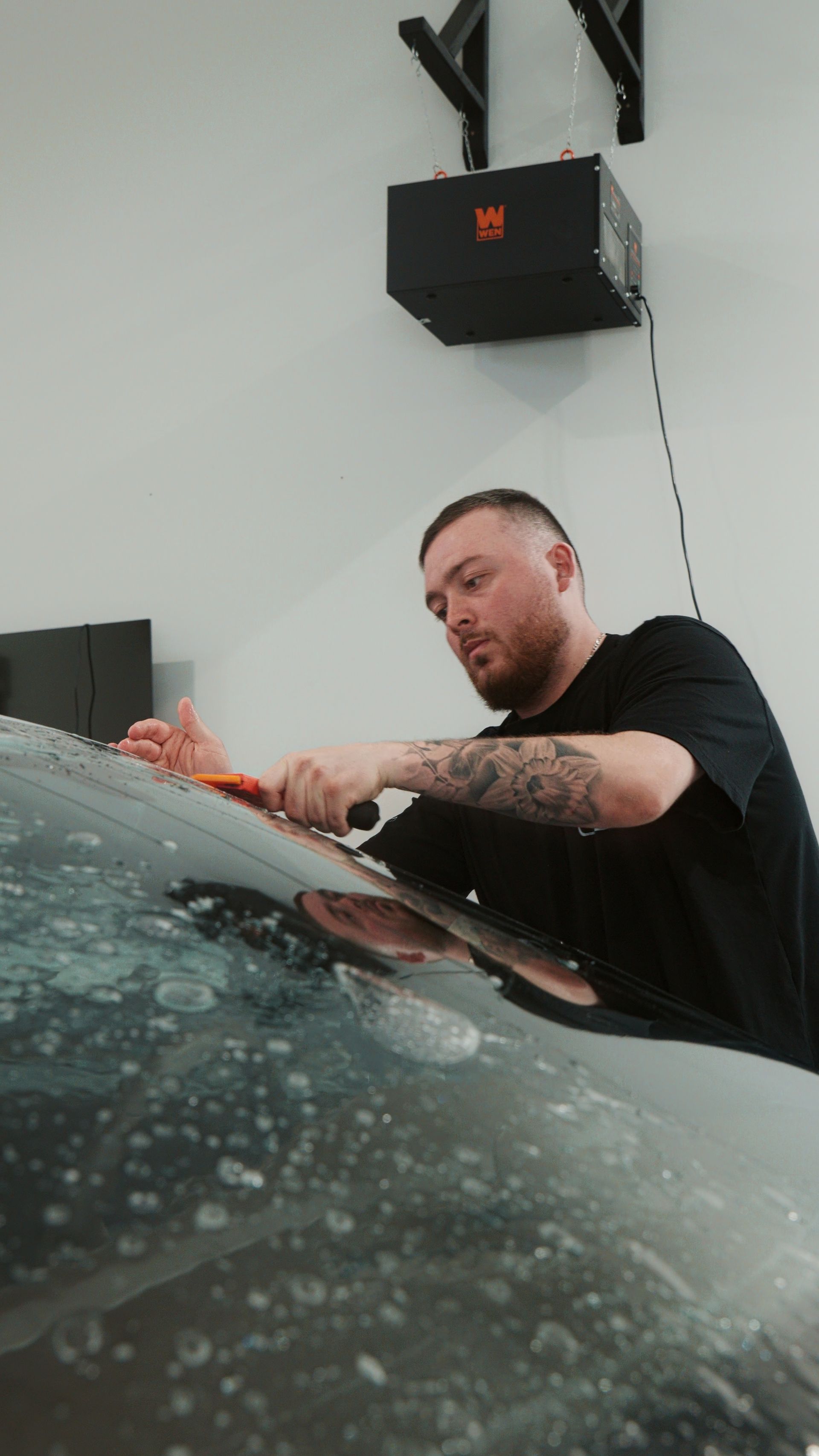 Man polishing a car windshield in a garage, with soap suds on the glass and a wall-mounted black box behind him