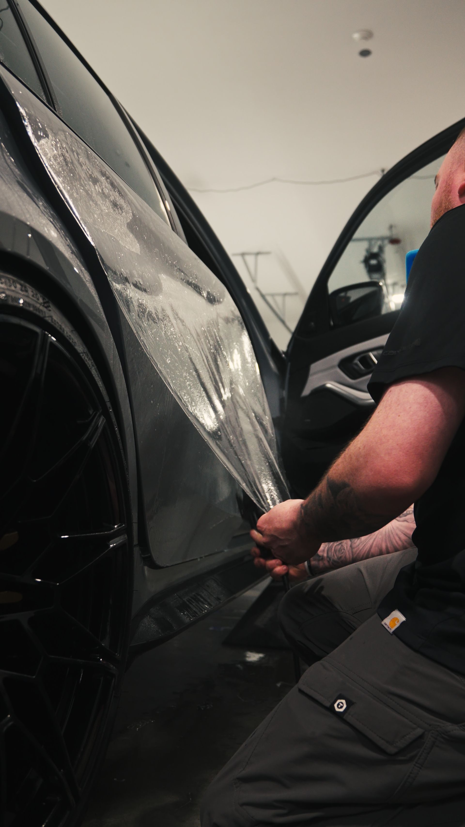 Person applying window tint to a car in a garage, holding a squeegee on the rear side window.