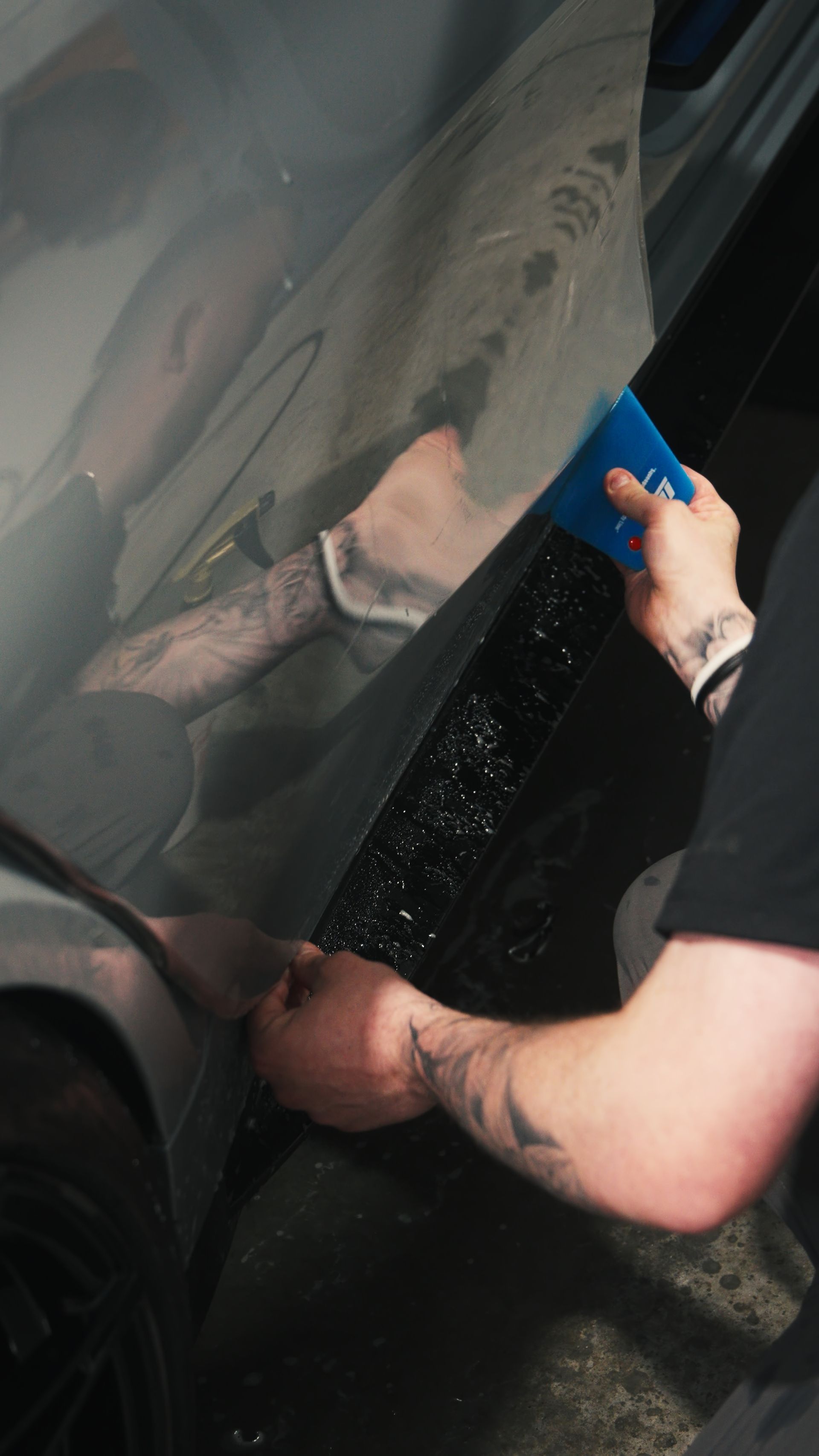 Person washing a black car door with a blue sponge, reflecting in the glossy paint.