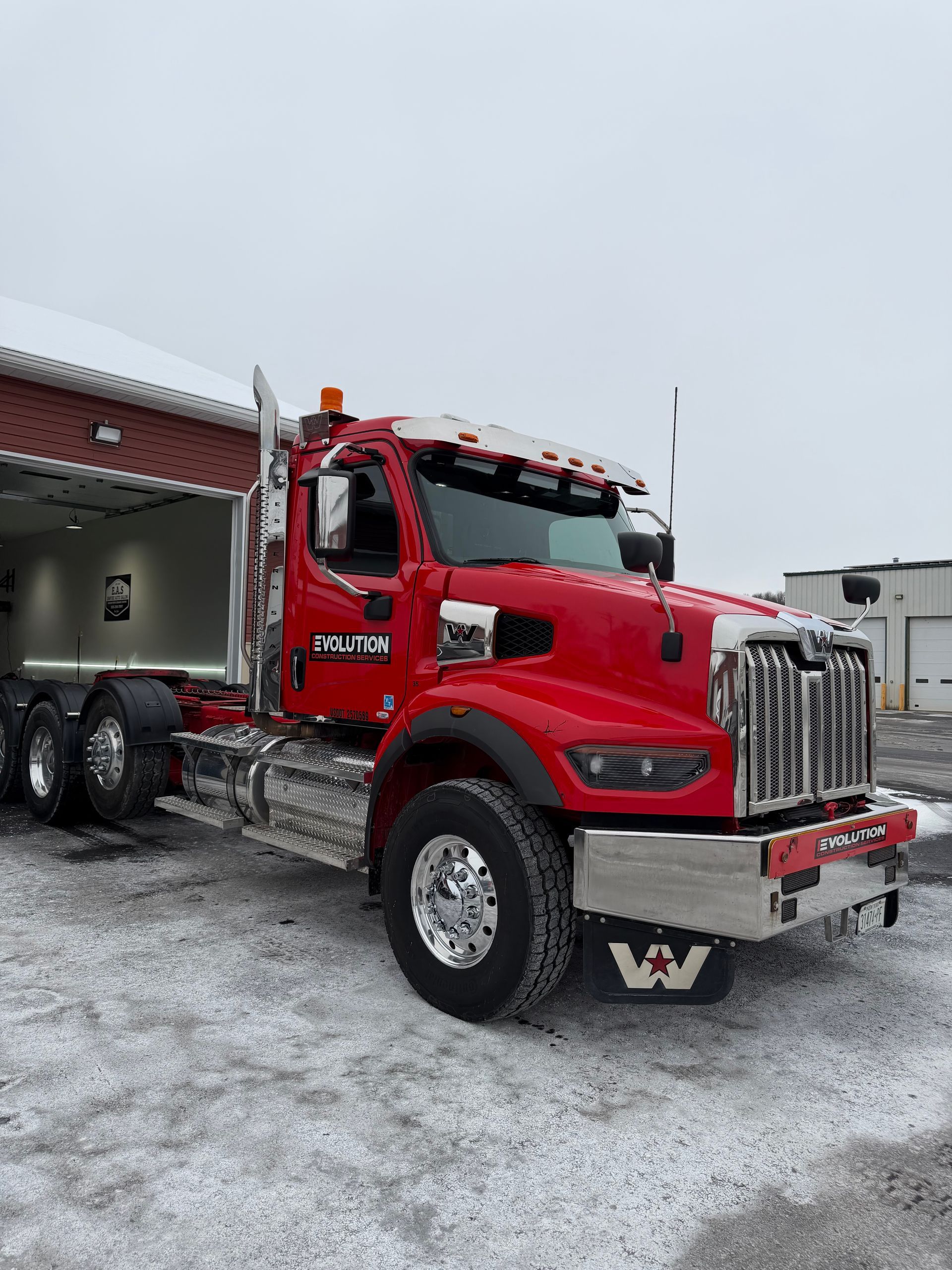 Red dump truck parked in snowy lot beside a building, front view with chrome grille and red cab