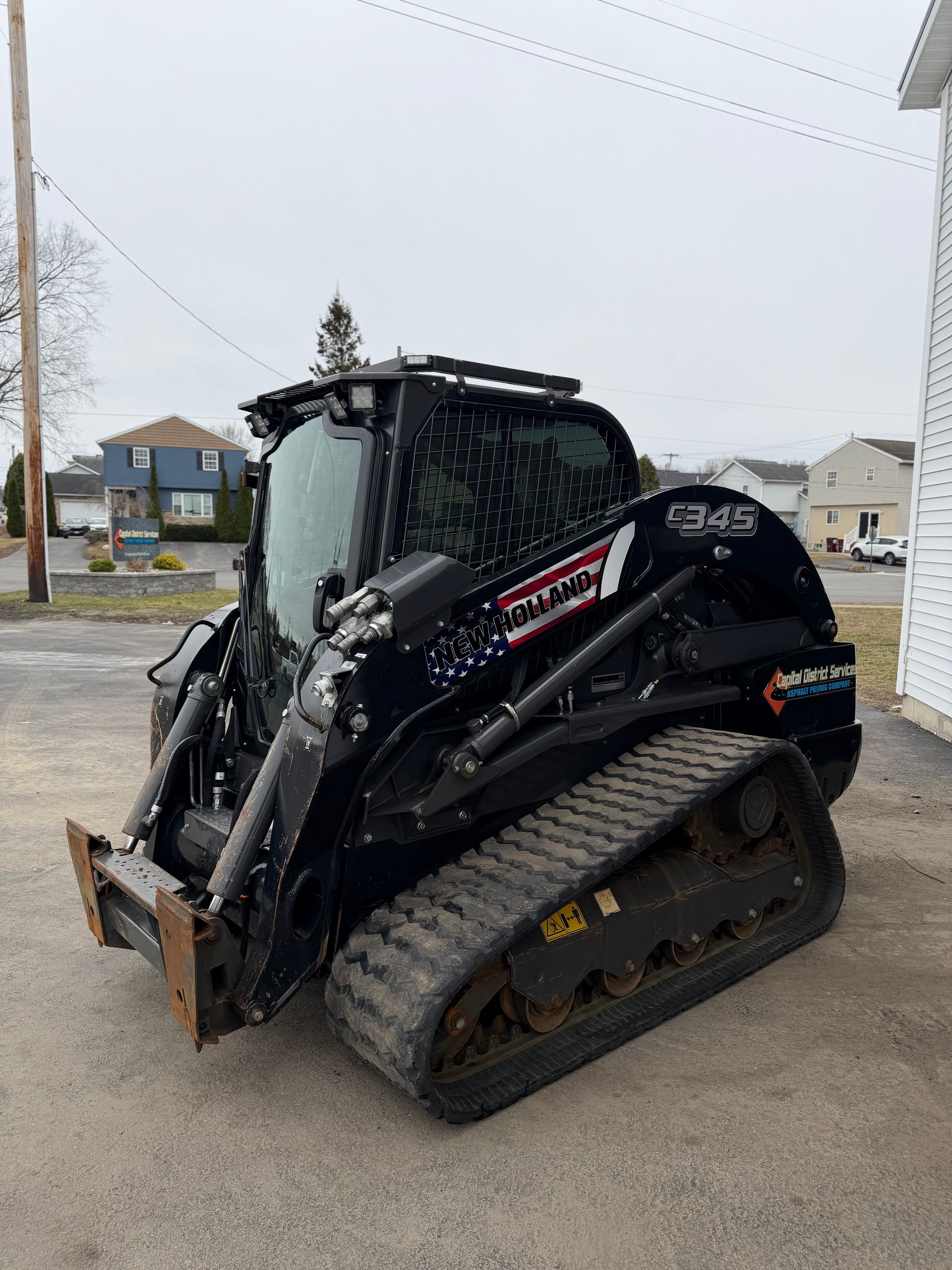 Black compact track loader with bucket parked on wet pavement in a snowy lot.