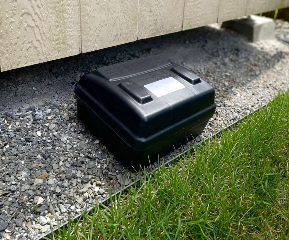 Orb Pest Solutions placed a black rodent bait box on gravel beside a beige building and grass.
