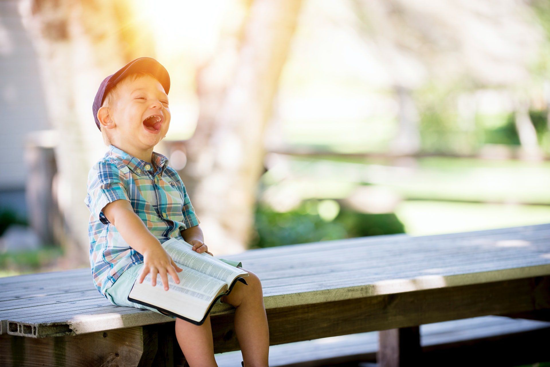 A young boy is sitting on a picnic table holding a bible and laughing.