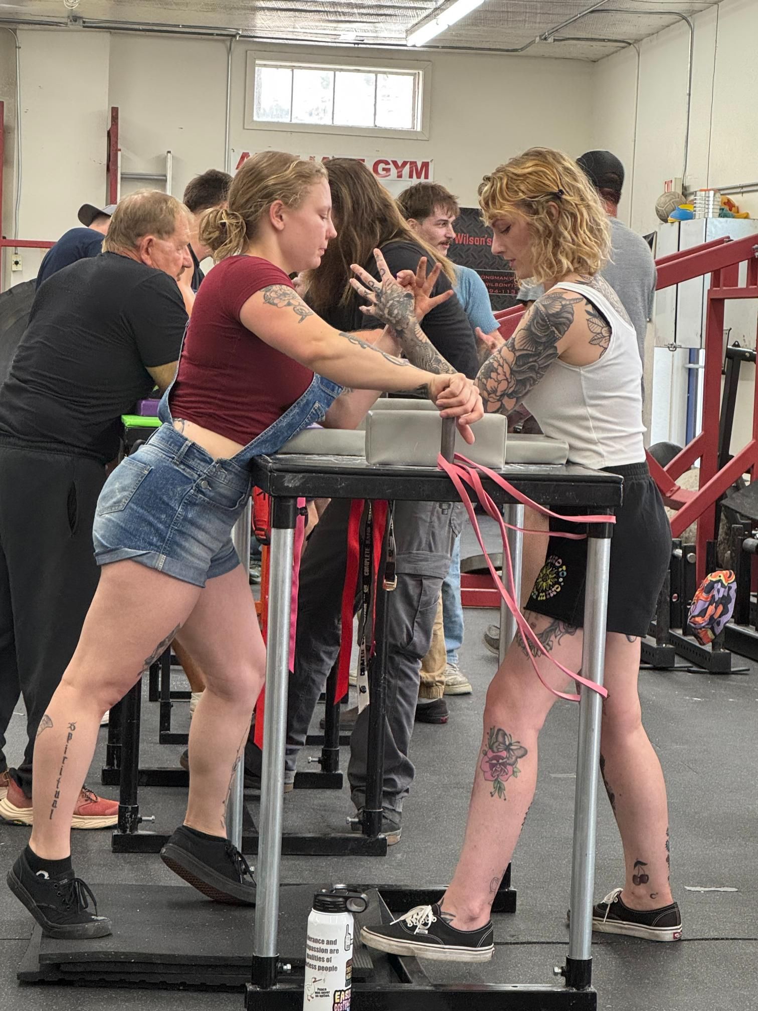 Two women arm wrestling at a gym. One wears overalls, the other a tank top, both have tattoos.