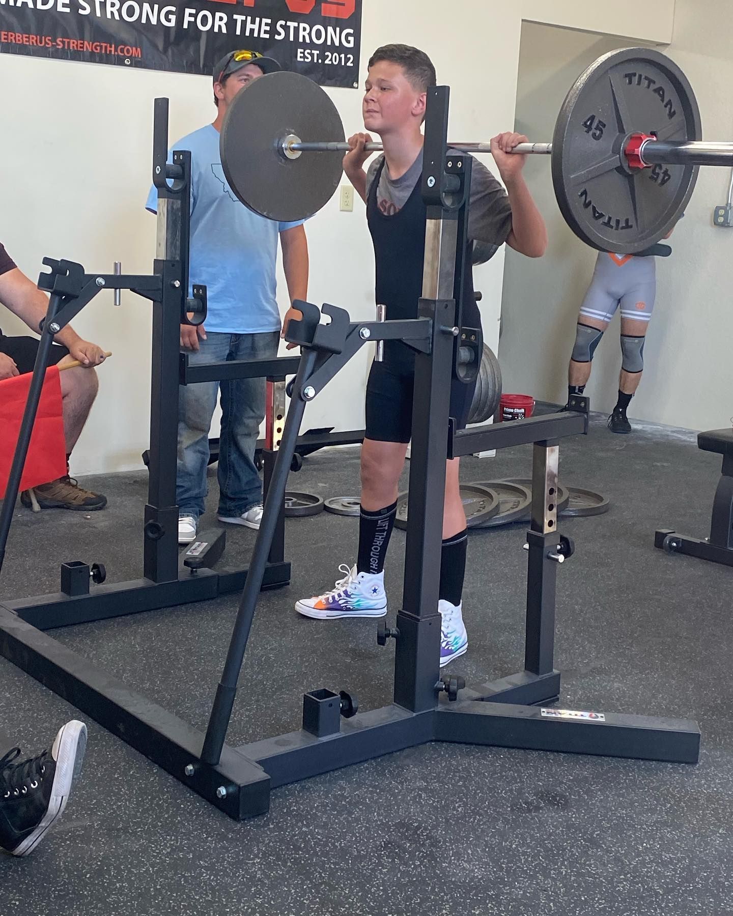 Young person squats with barbell at a gym, spotter behind, weights on bar.