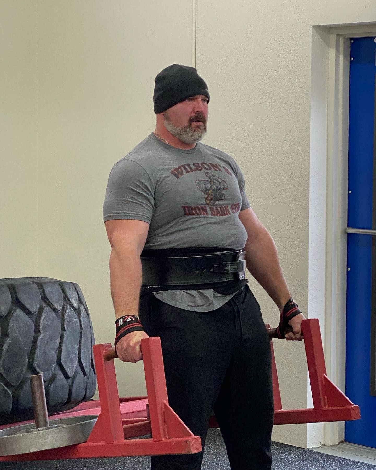 Man in a gym, lifting a weighted frame, wearing a lifting belt and beanie.