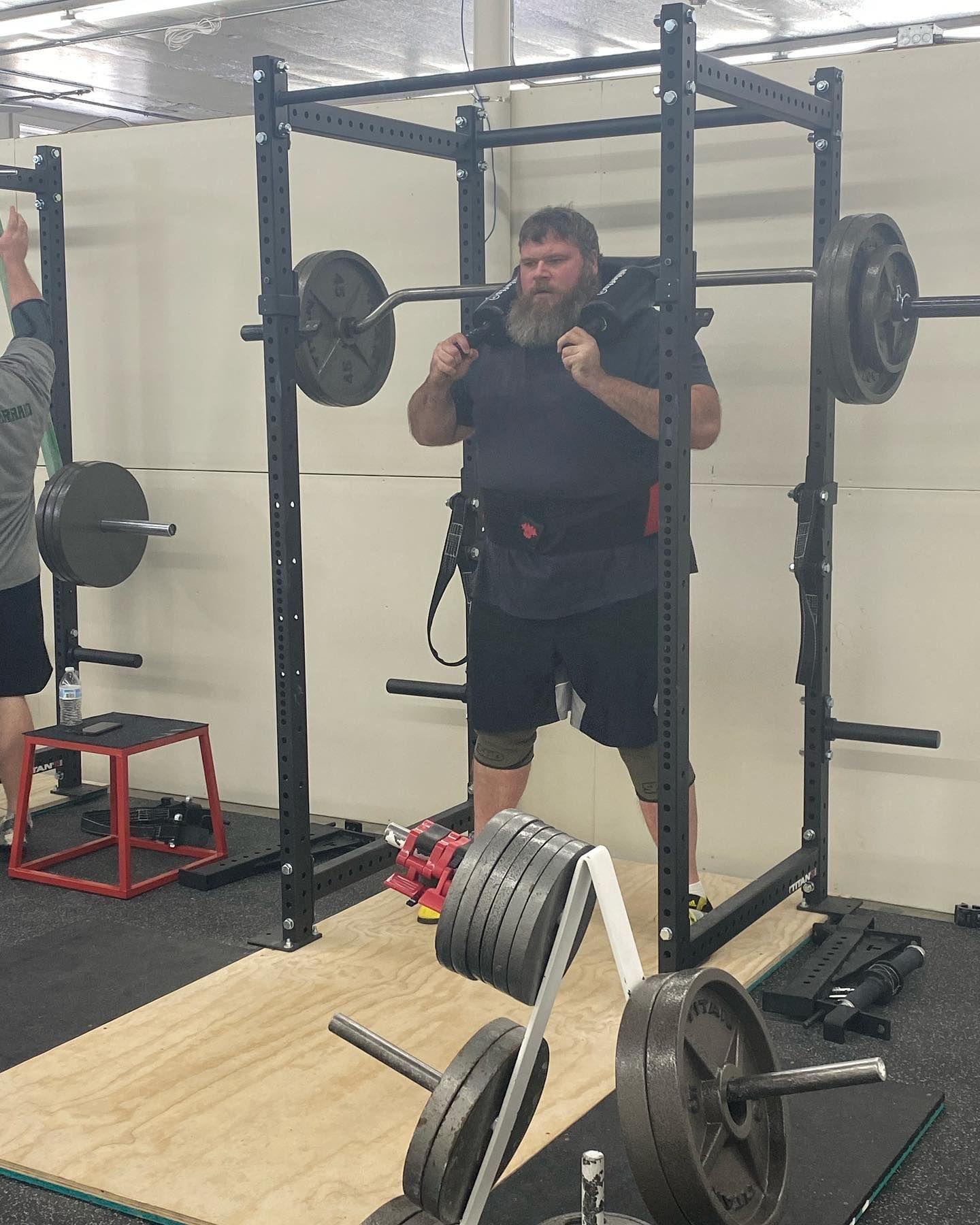 Man squats with barbell in a weightlifting rack. He wears a weight belt and kneepads.