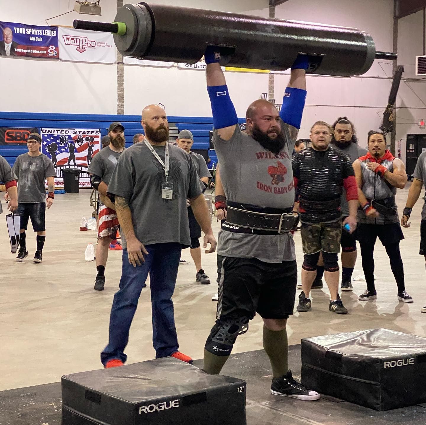 Man lifts heavy log overhead during a strongman competition. Others watch in a gym setting.