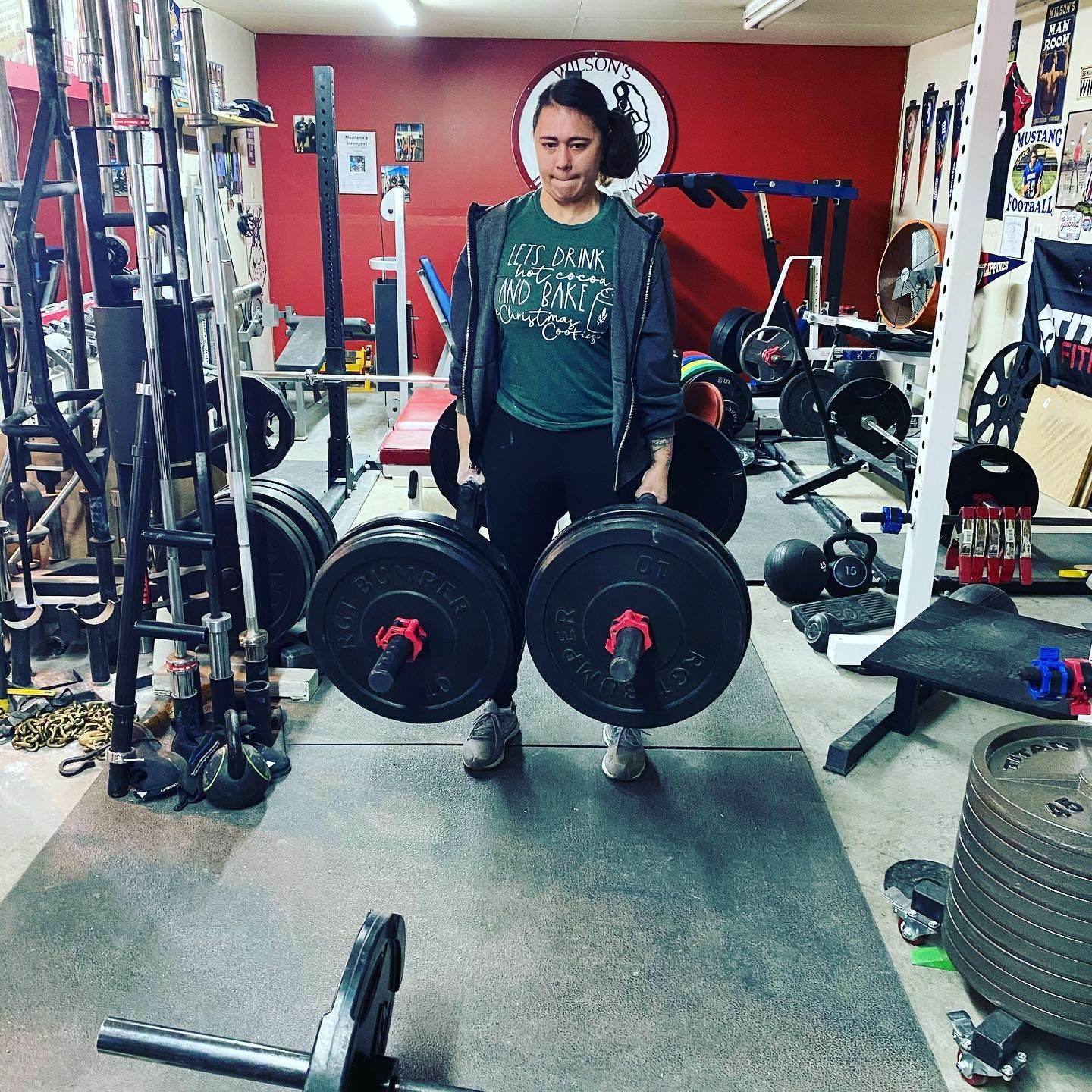 Woman in a home gym holding heavy dumbbells. Gym has red wall, weights, and various equipment.