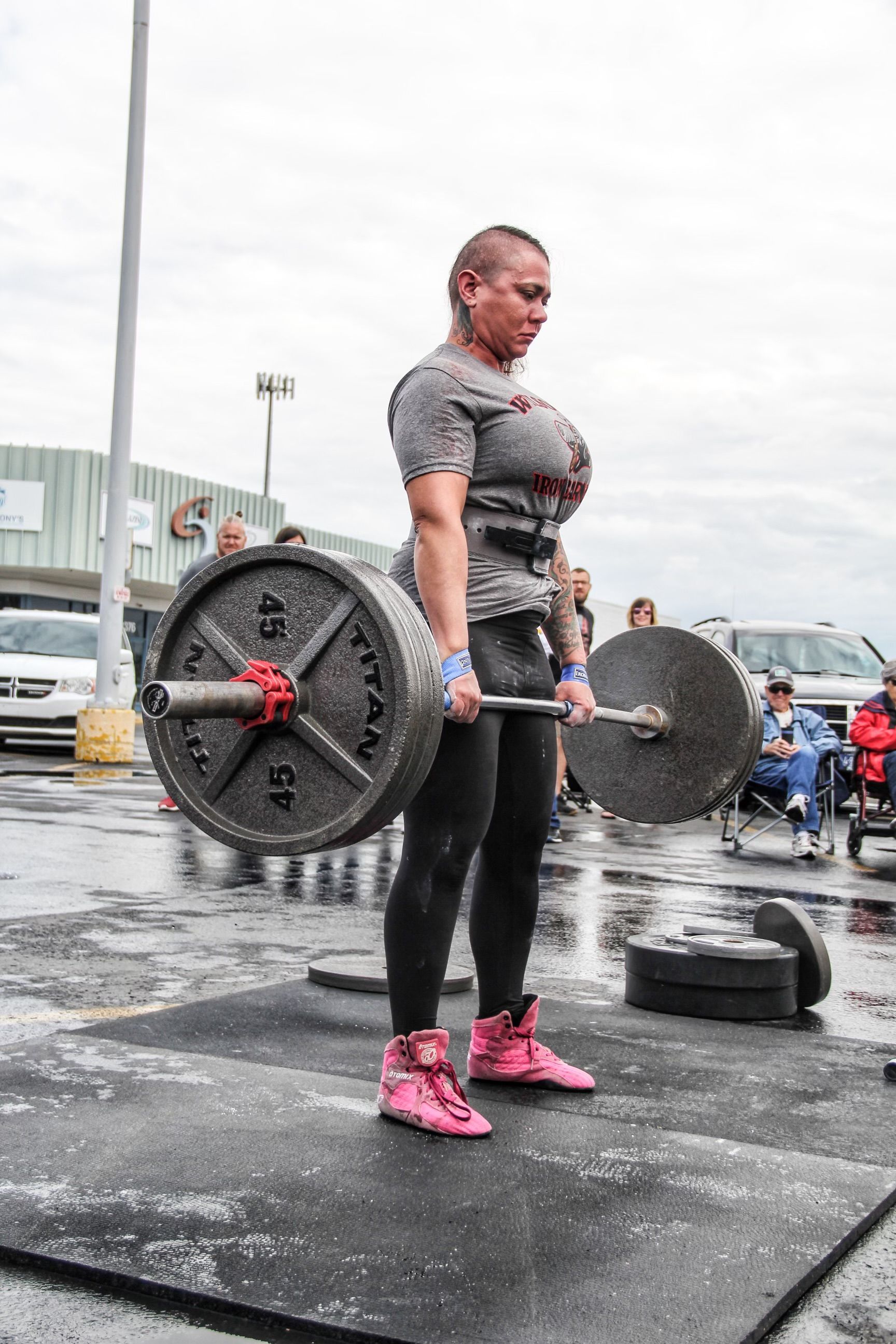 Woman in pink shoes deadlifting a heavy barbell outdoors on a rainy day.