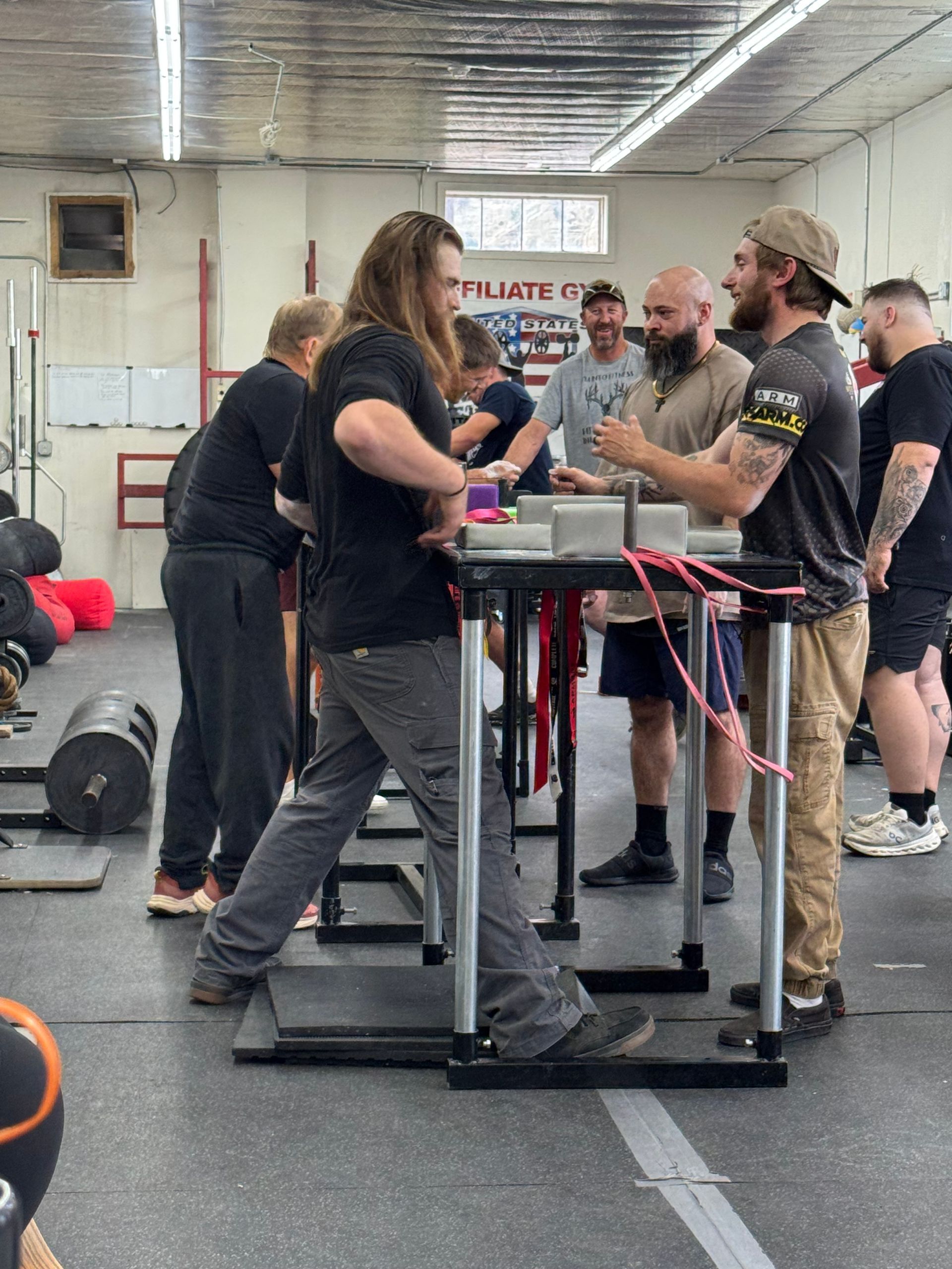 Arm wrestling match in a gym, two men at a table, others watching.