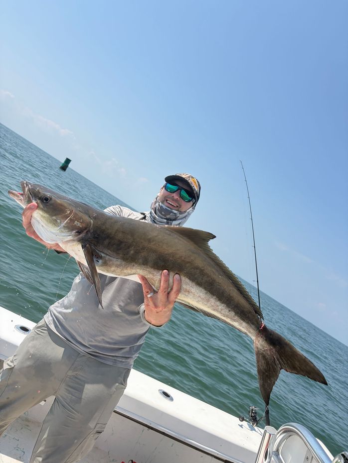 Man on a boat holding a large striped bass. He's wearing sunglasses and a jacket, with the ocean and engine visible.