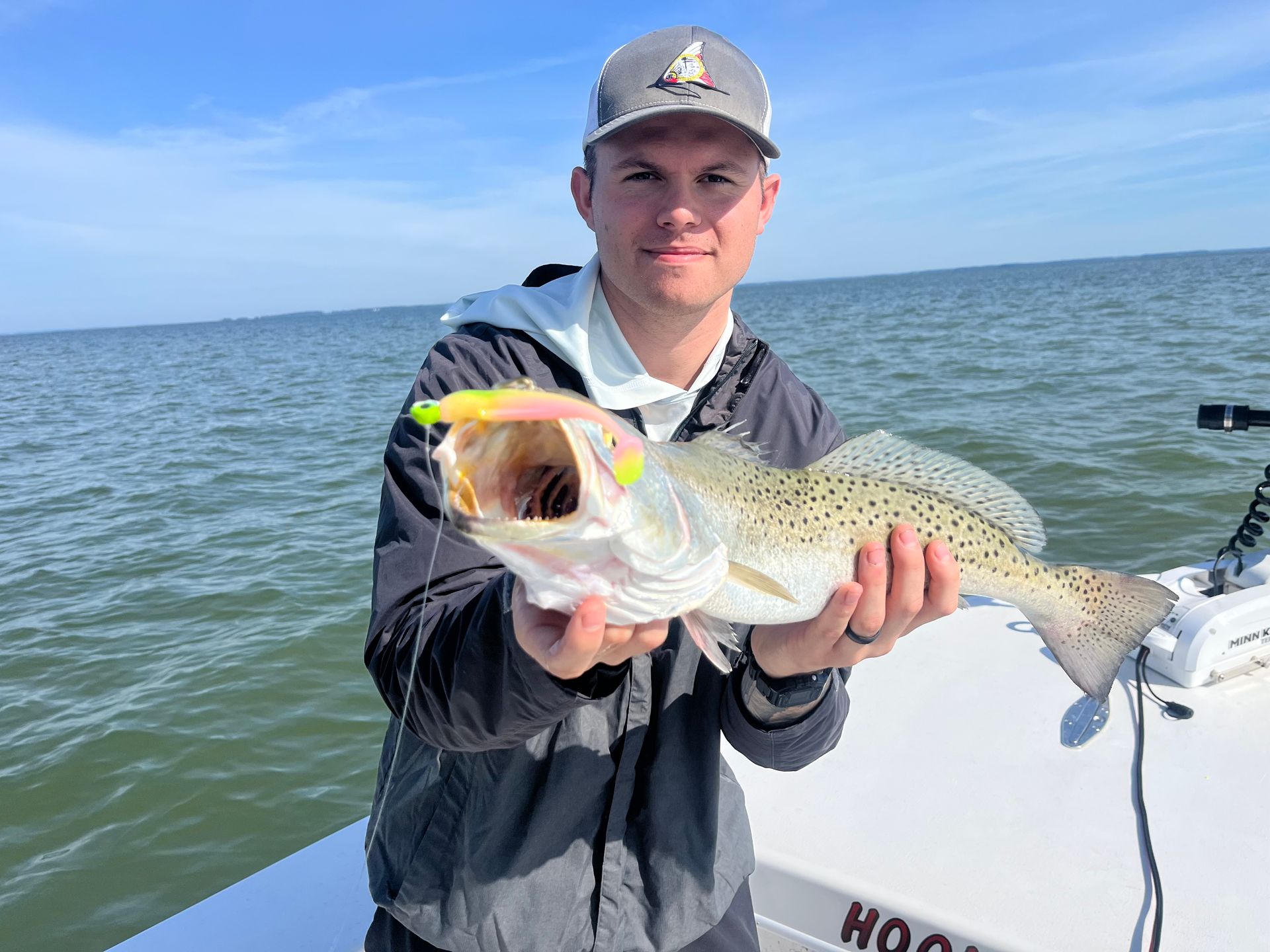 Man holding a spotted sea trout on a boat, the fish has a fishing lure in its mouth.