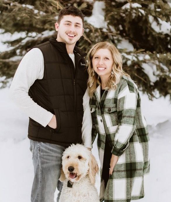 A man and a woman are posing for a picture with a dog in the snow.