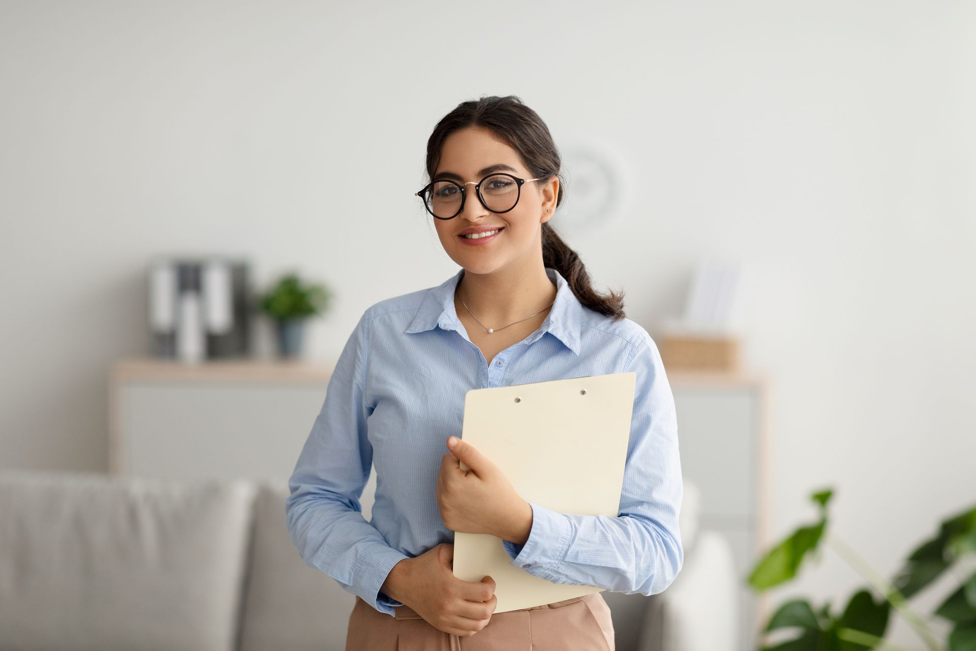 Woman in glasses smiling, holding a folder, in a home office setting. Woman in glasses smiling, holding a folder, in a home office setting.