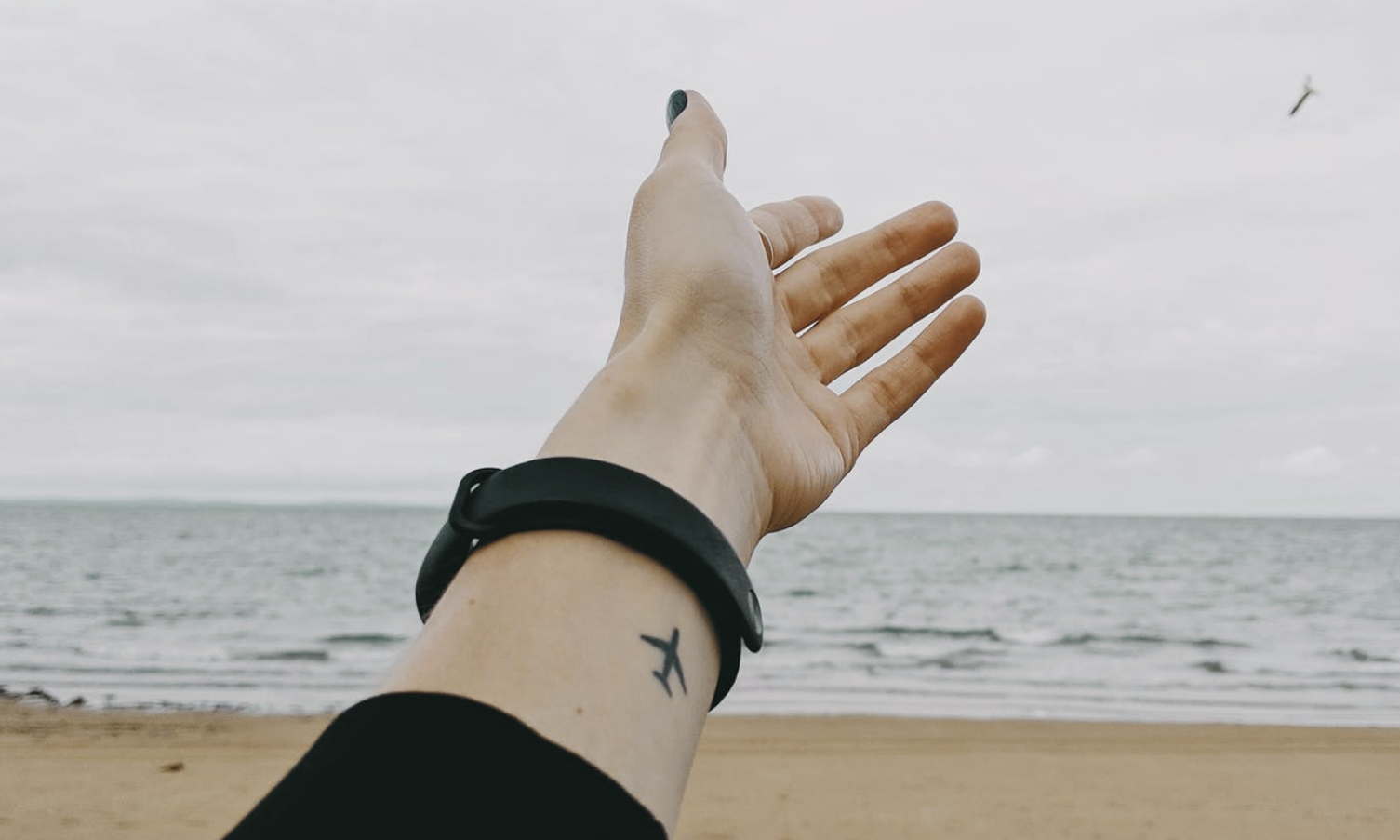 Arm outstretched towards an airplane, showing an airplane tattoo. Beach and sea in background.