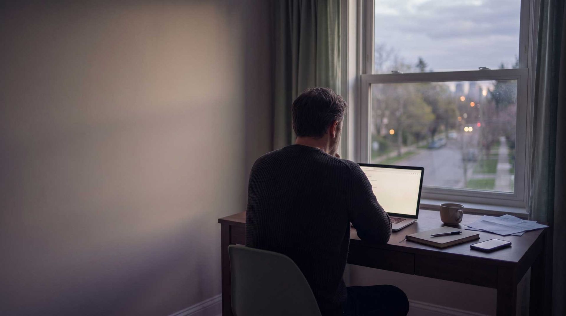 Person seated at a desk by a window, working on a laptop in a softly lit room.