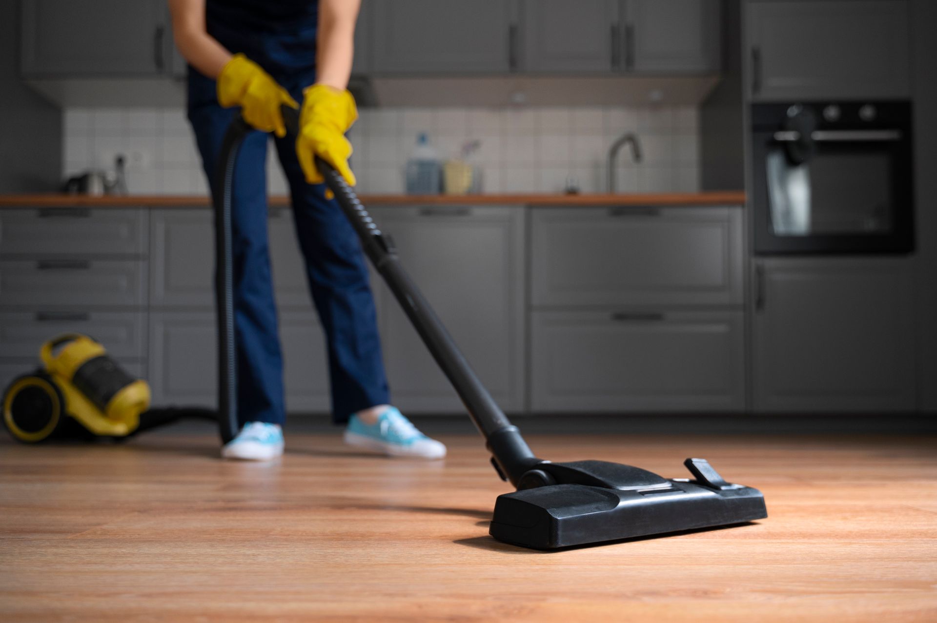 Person vacuuming a hardwood floor in a kitchen with a black vacuum and yellow gloves