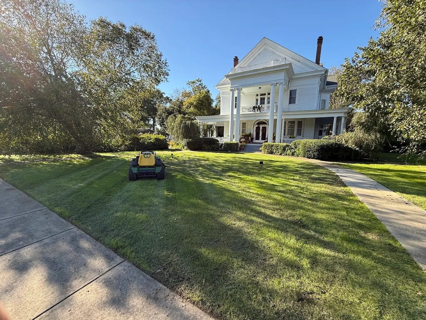 White historic mansion with columns on a sunny lawn, framed by trees and a sidewalk in the foreground