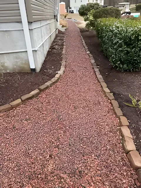 Red gravel side path beside a house, bordered by tan stones and a hedge.