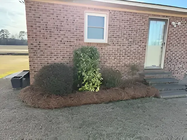 Brick building exterior with small shrubs, a window, a door, and steps beside a gravel lot