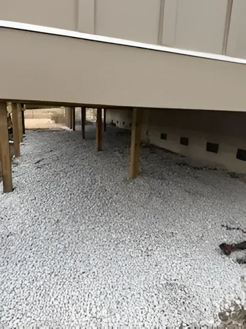 Gravel-covered crawlspace beneath a beige raised deck and mobile home, with wooden support posts.