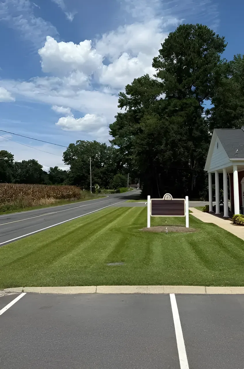 Roadside sign on a manicured lawn beside a building, with trees and cloudy sky in the background.