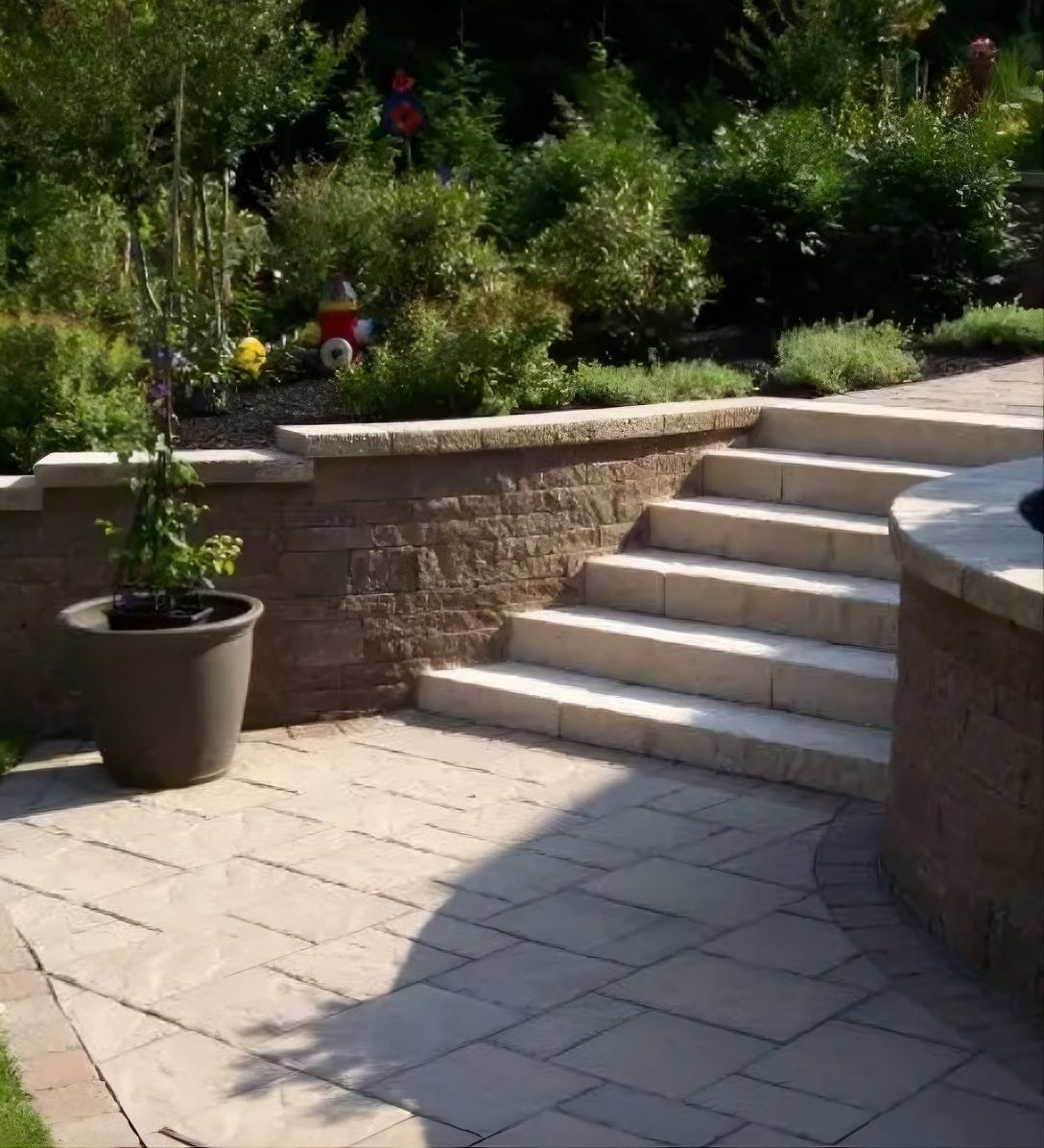 Sunny garden patio with stone steps, paver walkway, and potted plants beside raised flower beds