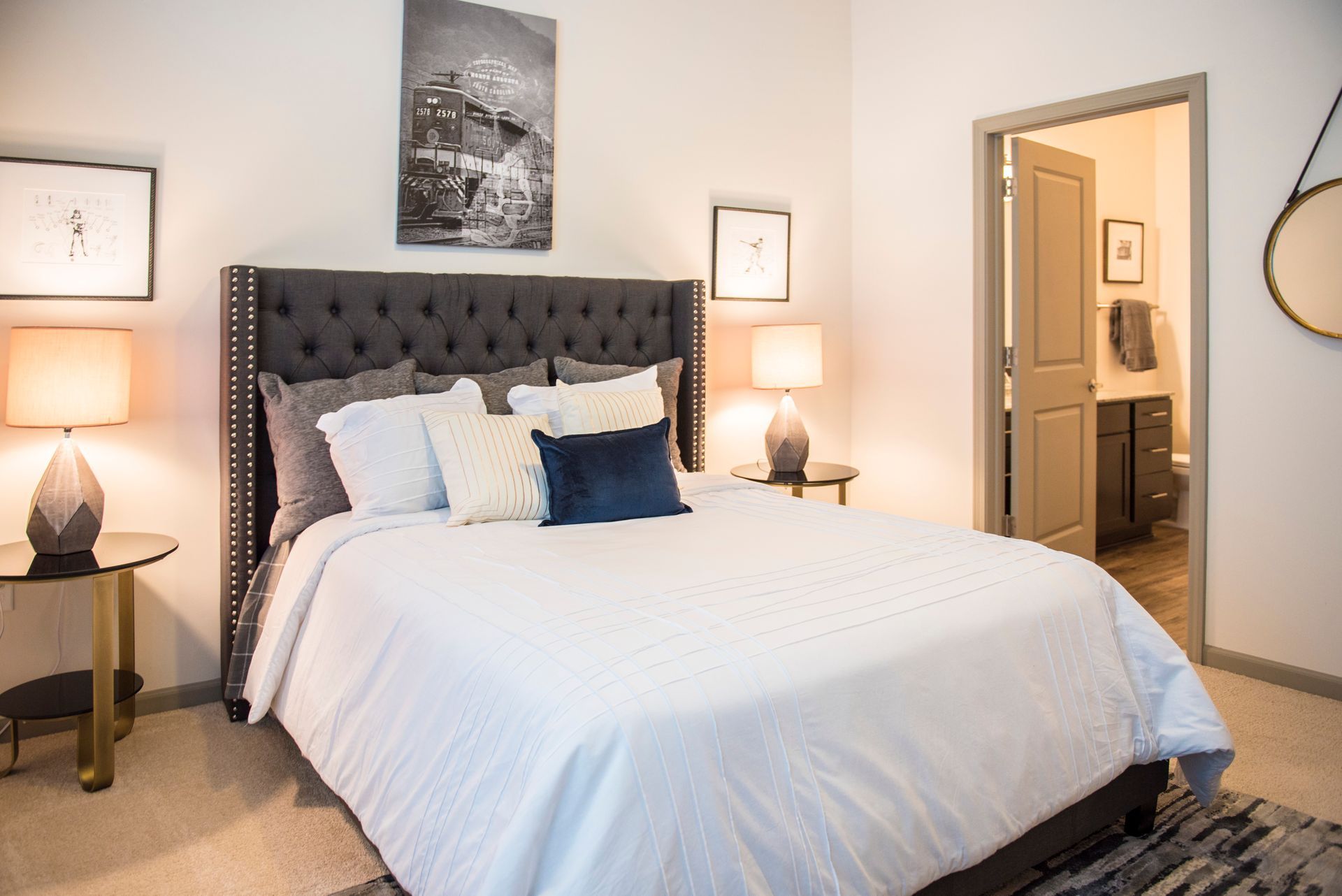 Bedroom with a gray upholstered headboard, lamps on side tables, artwork, and an open bathroom door at Ironwood Apartment Homes.