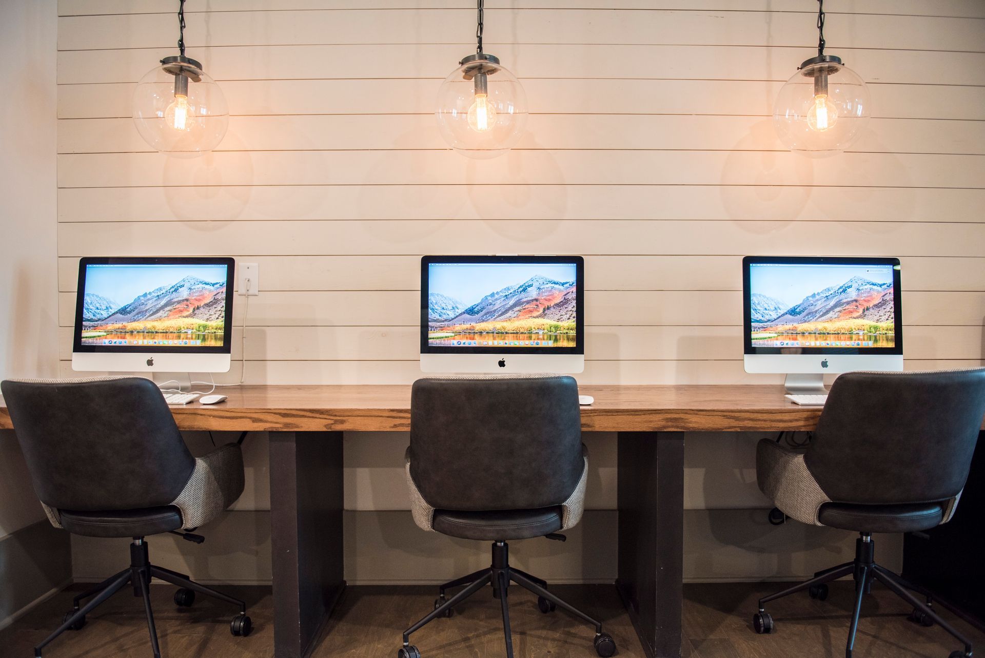 Three desktop computers on a wooden desk in a modern office, with three chairs and pendant lights at Ironwood Apartment Homes.