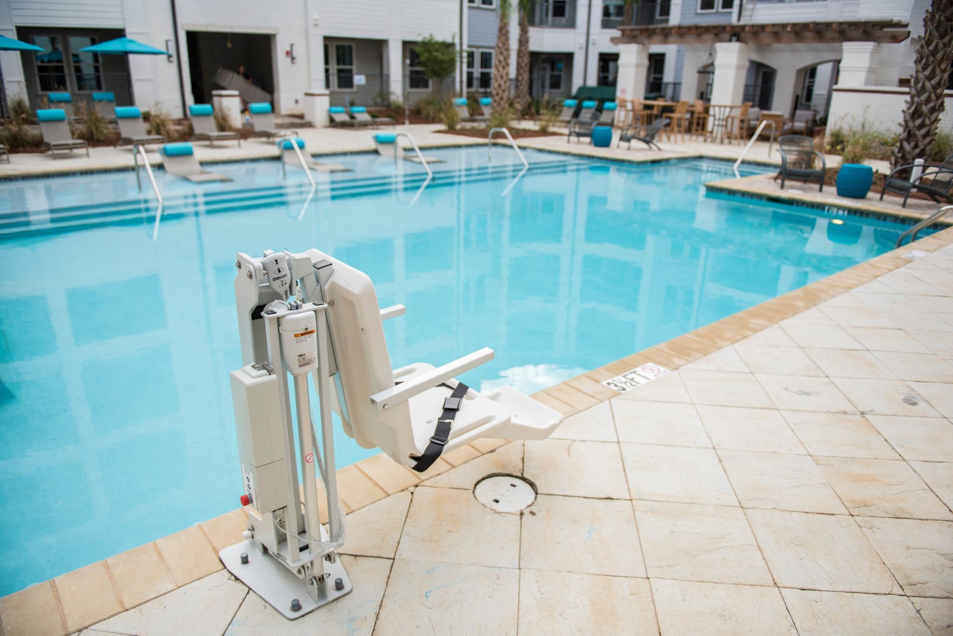Poolside chair lift; cream-colored chair, blue pool, light stone tiles, apartment building in background at Ironwood Apartment Homes.