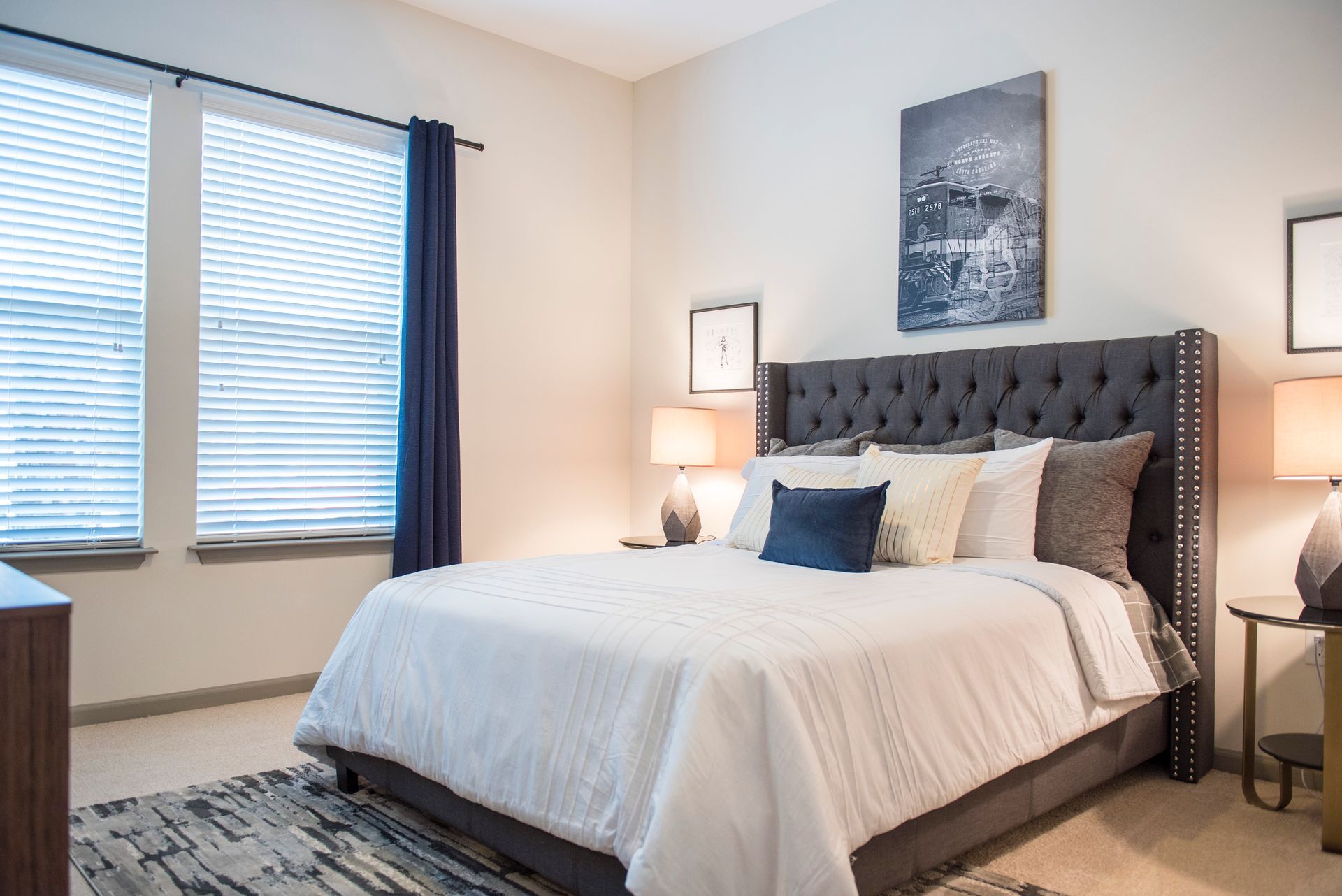 Bedroom with a bed, two windows, art, and bedside tables; beige walls, dark blue accents at Ironwood Apartment Homes.