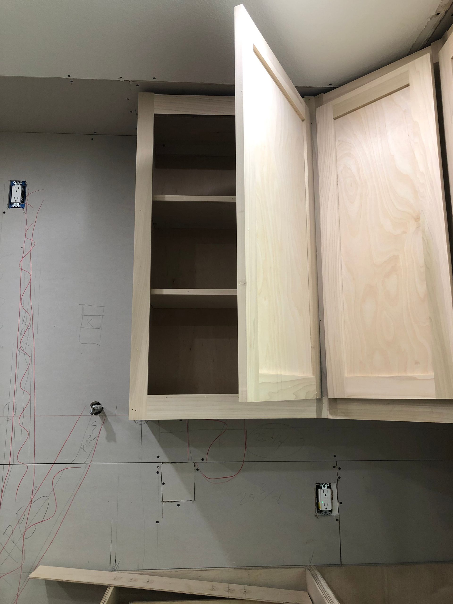 An open unfinished wooden kitchen cabinet mounted on a grey drywall wall with an electrical outlet visible below.