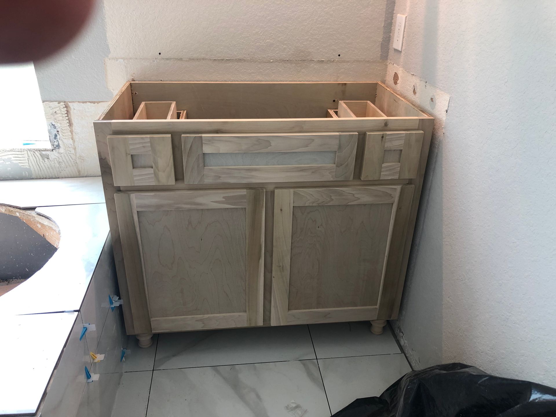 An unfinished wooden bathroom vanity cabinet installed between a wall and a tiled tub surround.