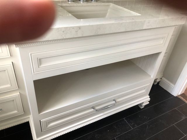 A white bathroom vanity with an open center shelf, a bottom drawer, and a light-colored marble countertop.