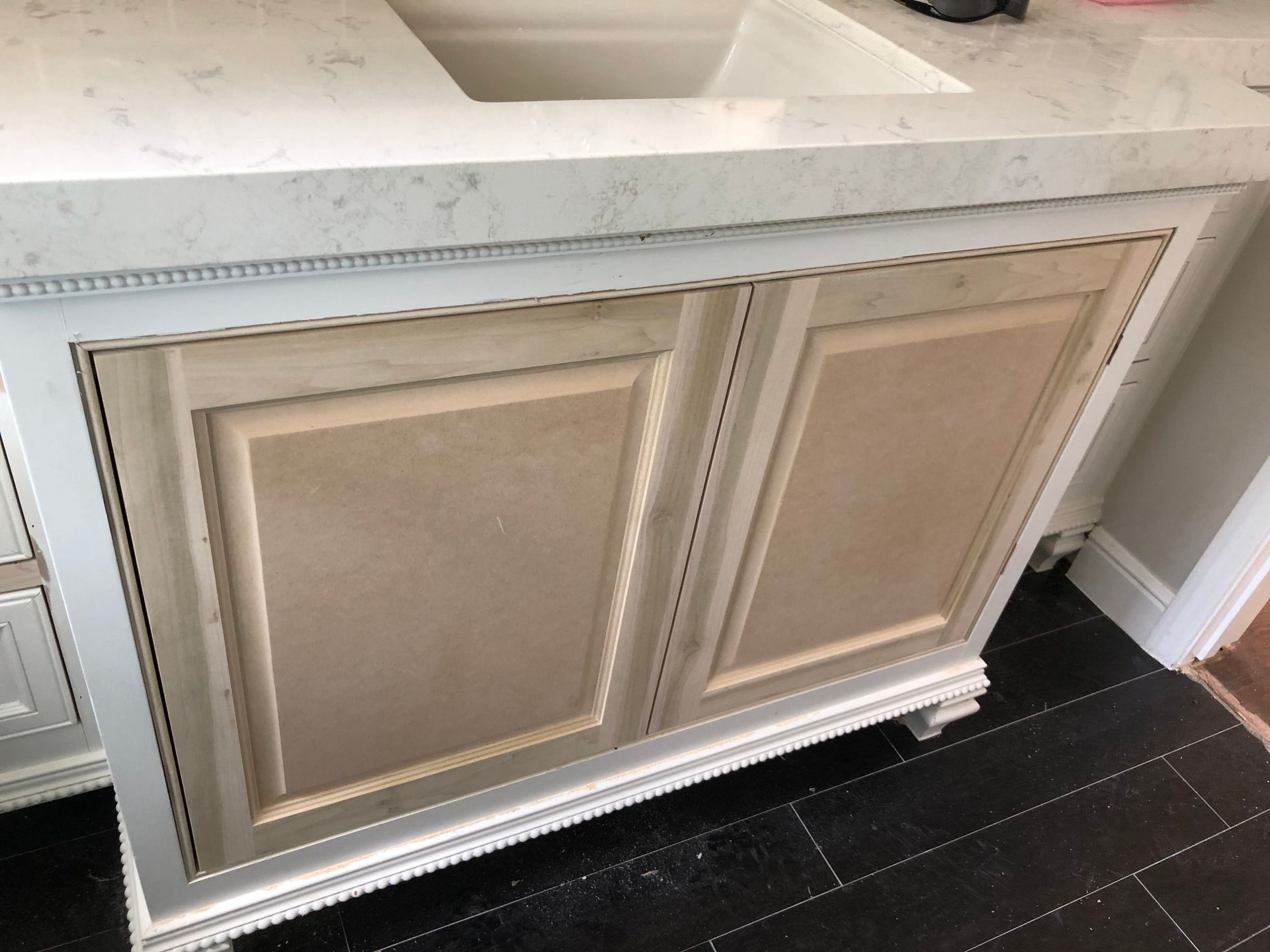 A close-up of a white bathroom vanity with unfinished wood cabinet doors, a quartz countertop, and black tiled flooring.