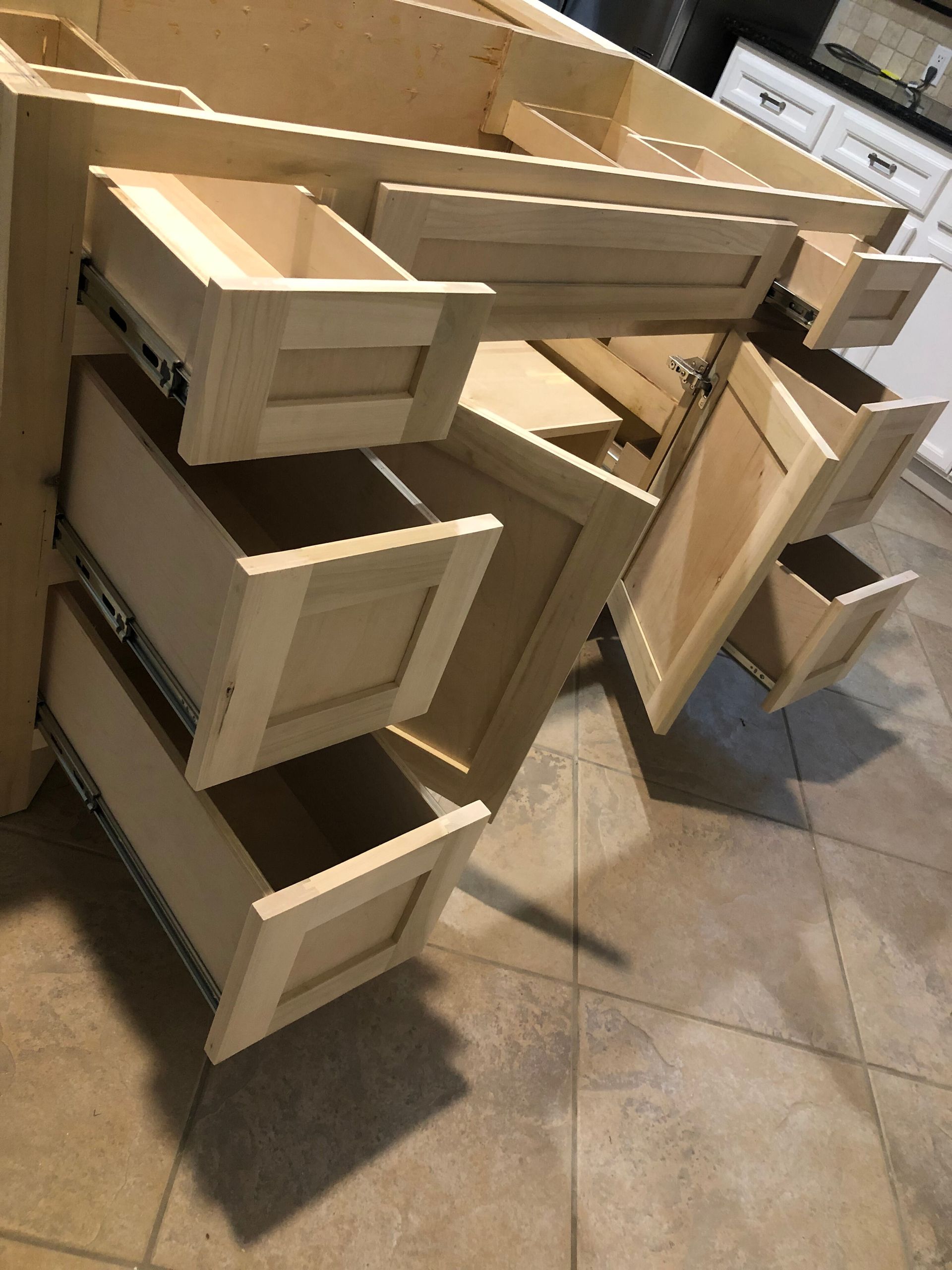 An unfinished, light wood bathroom vanity cabinet with multiple open drawers and a door on a tiled floor.