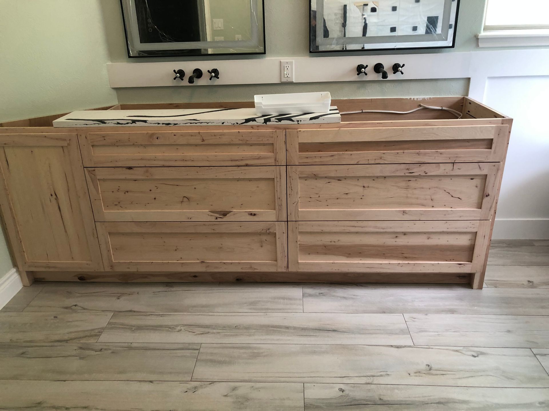 A bathroom vanity with unfinished wood drawers and a white countertop, featuring two mirrors and wall-mounted faucets.