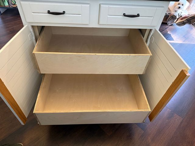 A white kitchen cabinet with two open doors, revealing two stacked wooden pull-out drawers, sitting on a wooden floor.