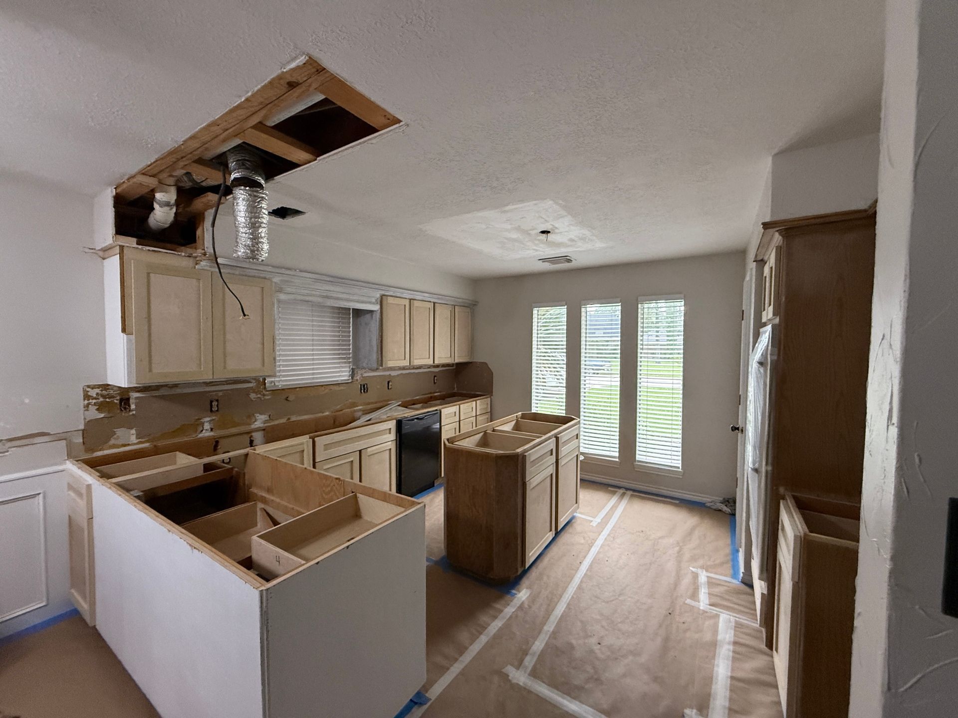 An under-construction kitchen with light wood cabinets, an island, and exposed ceiling framing in a brightly lit room.