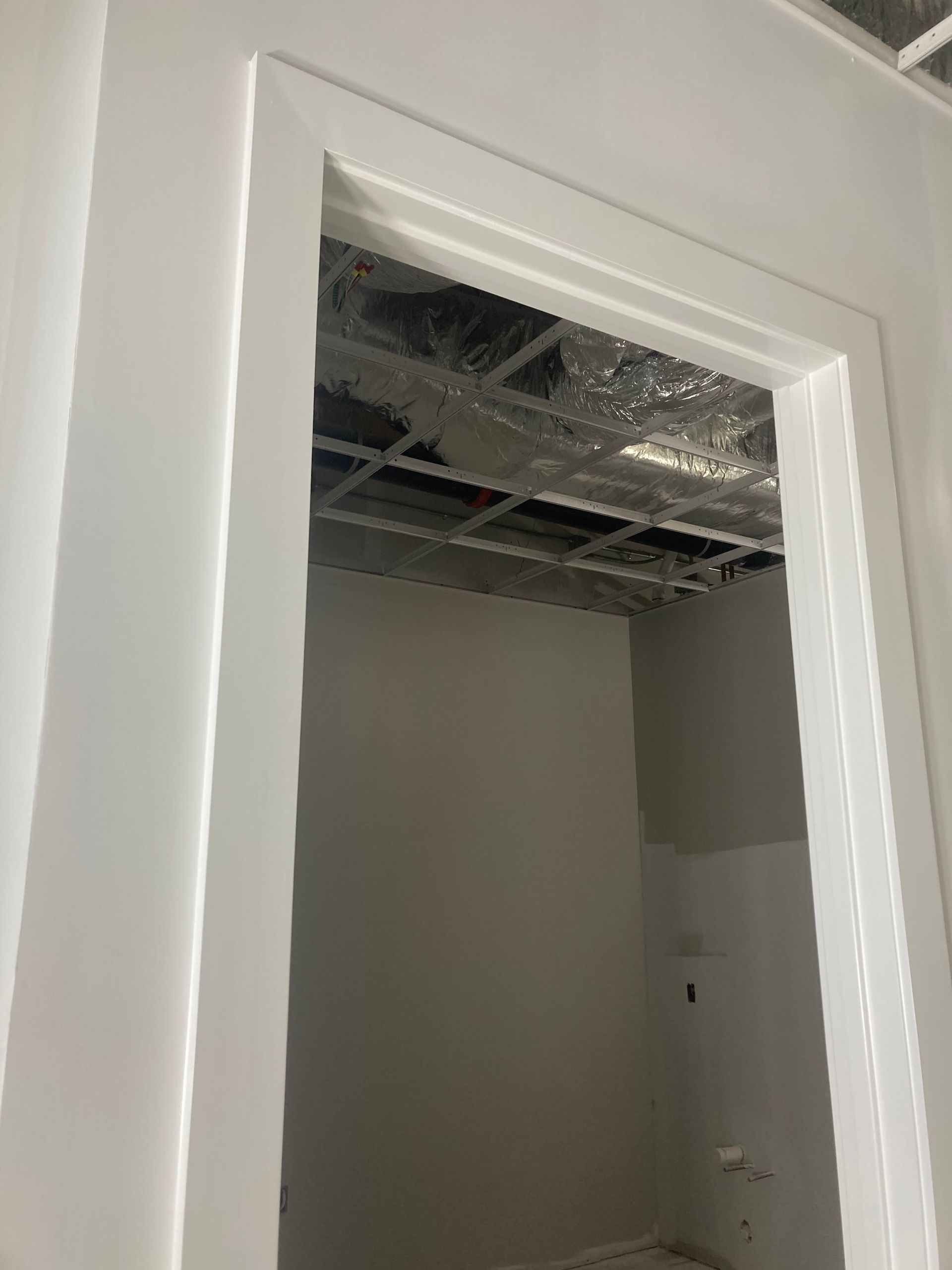 A white-trimmed door frame opens into an unfinished room with an exposed metal ceiling grid and partially painted walls.