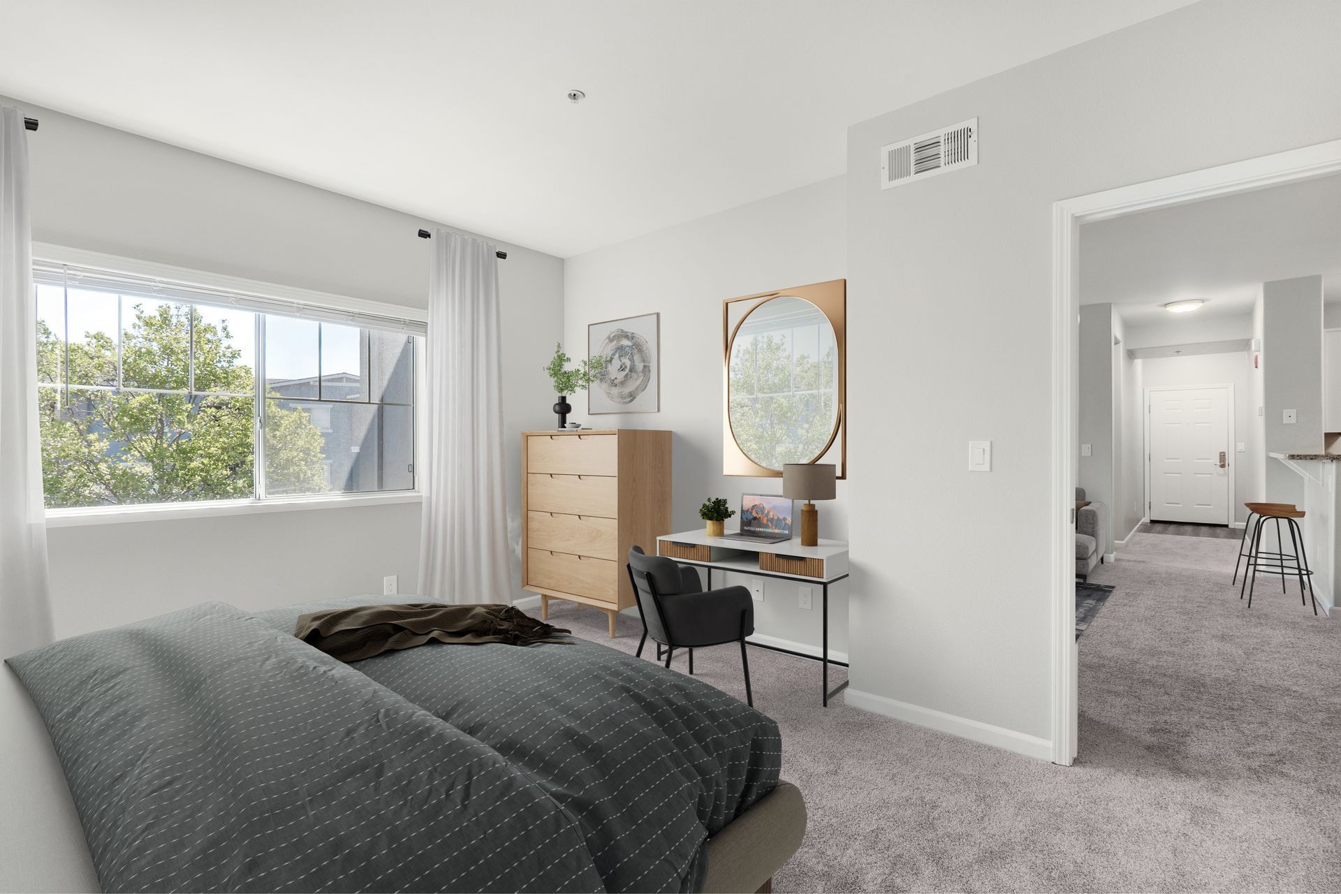 Bright bedroom with gray bedding, dresser, vanity, and open doorway to a carpeted hall