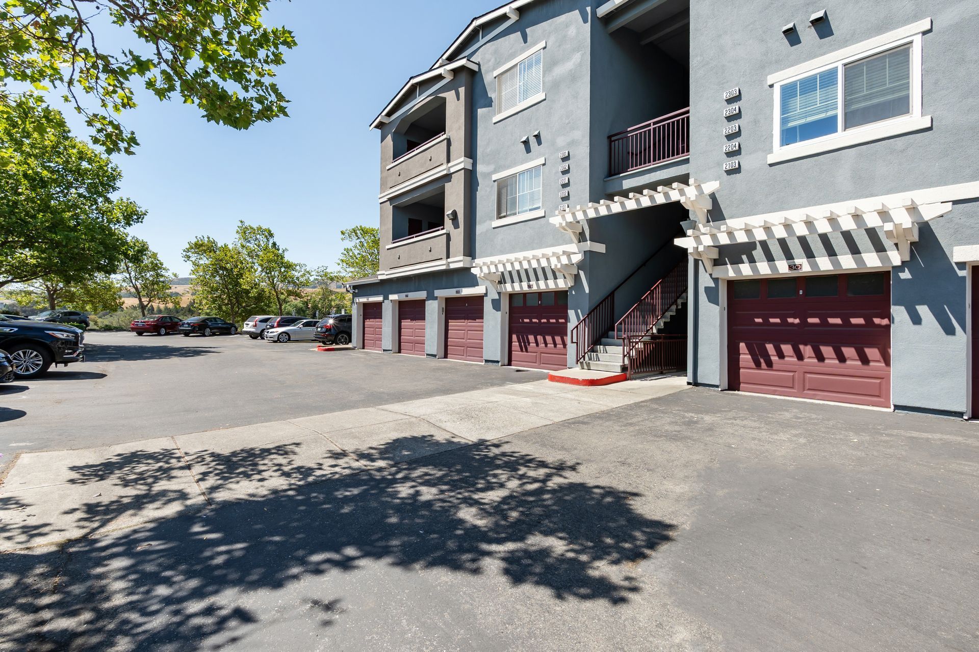 Apartment building with garage doors and striped awnings beside a parking lot on a sunny day