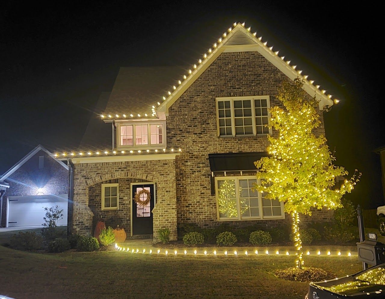 Brick house at night outlined in warm white lights, with a lit tree in the front yard.