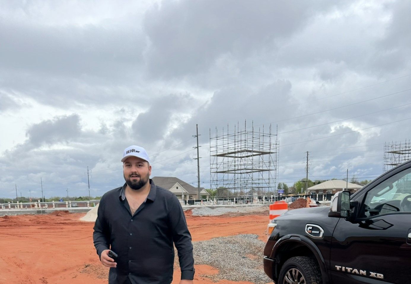 Man standing on a red dirt construction site beside a black pickup truck under cloudy skies