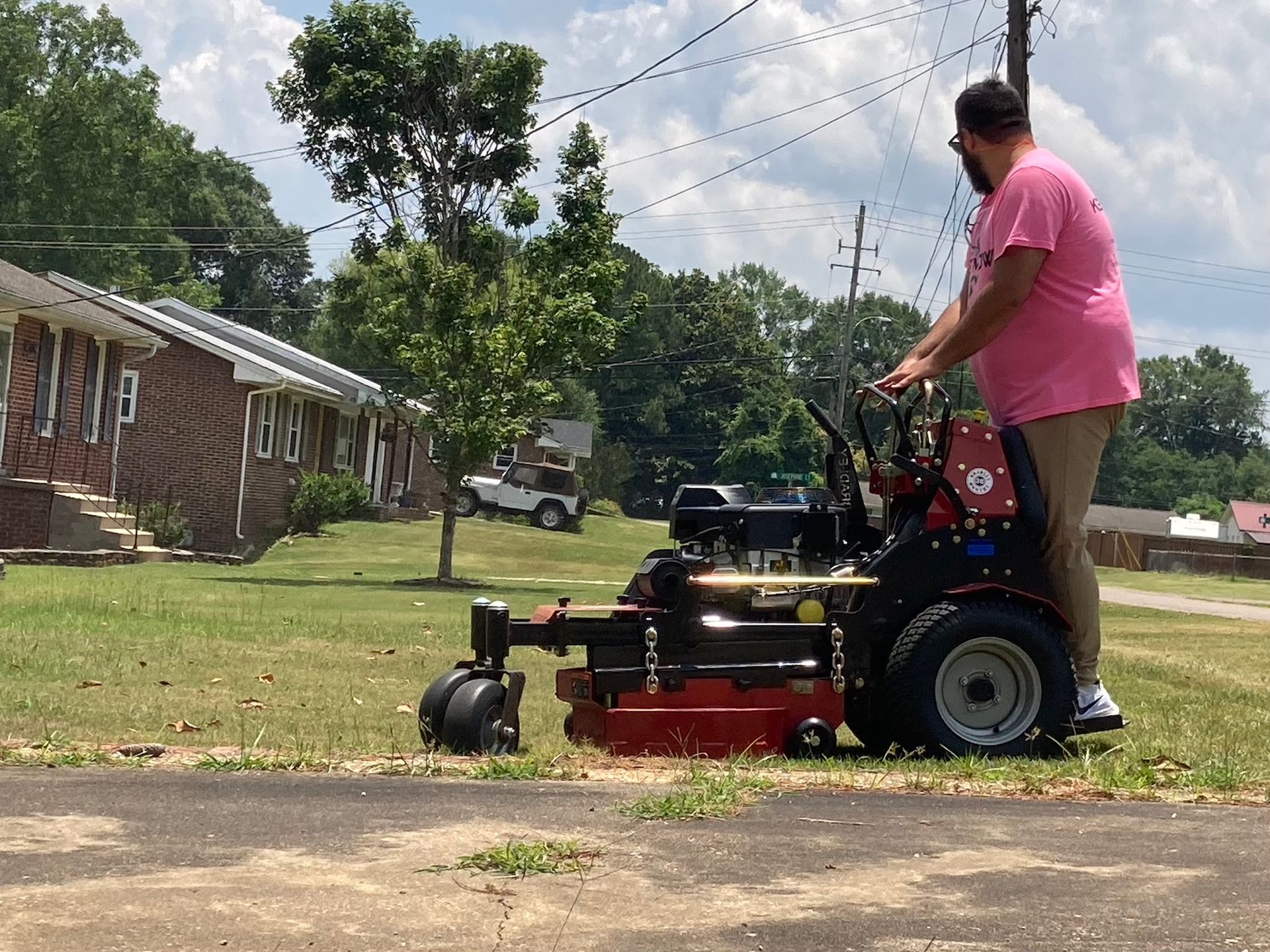 Person riding a red-and-black zero-turn mower on a grassy yard beside a brick house