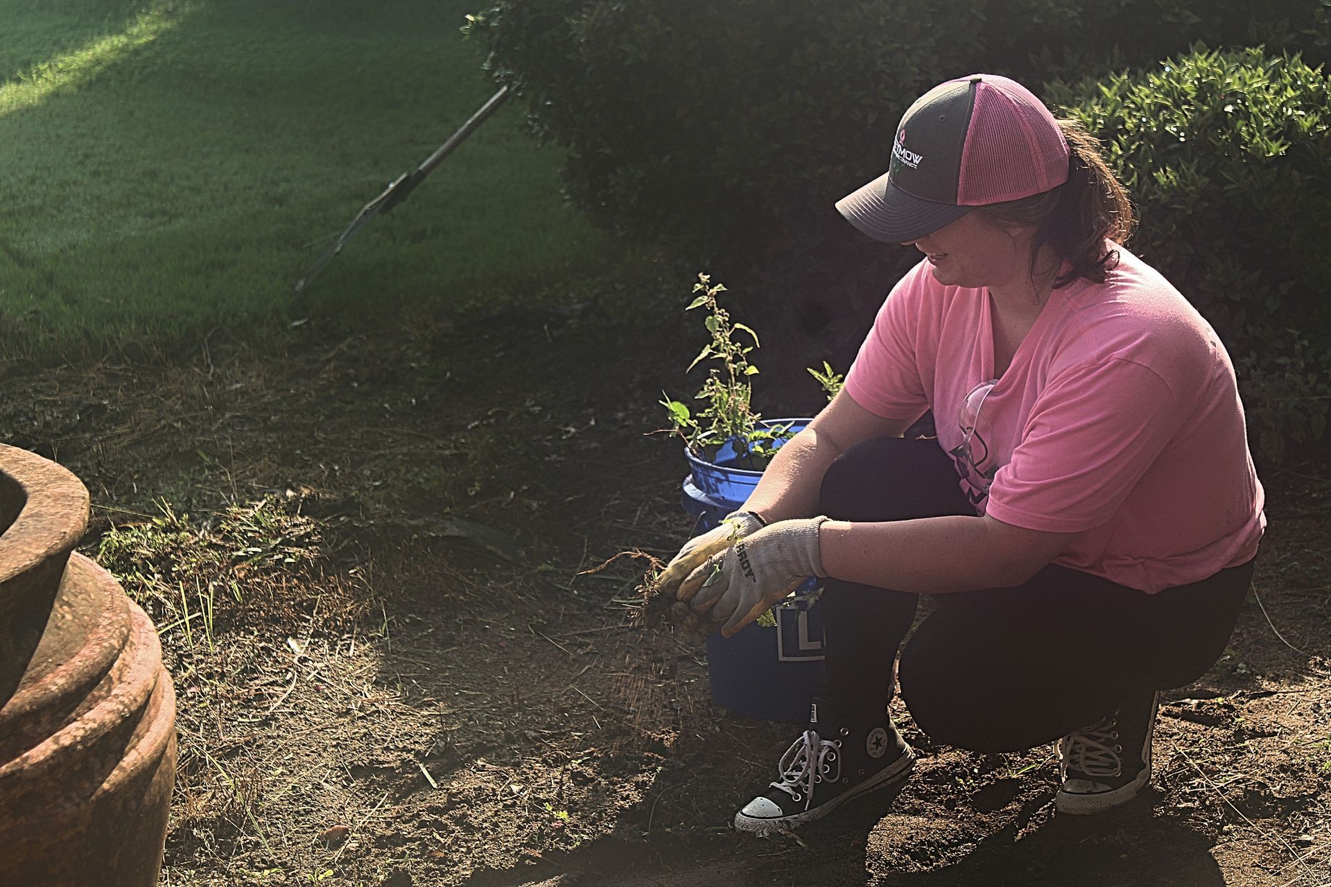 Person crouching outdoors beside a small plant, wearing a pink shirt and cap, near a pond and stone steps.