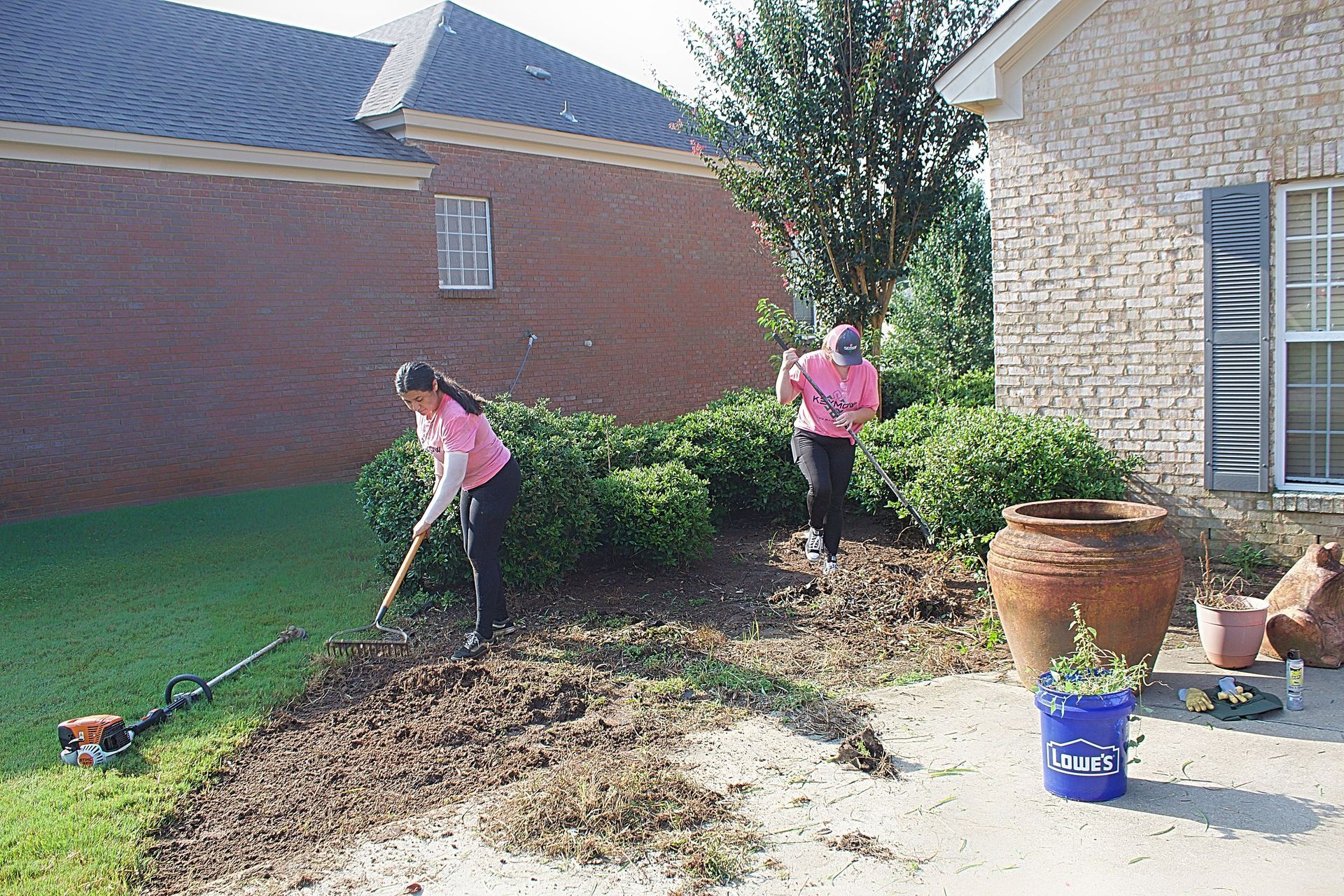 Two people in pink shirts working in a garden beside a brick house, raking and trimming bushes.